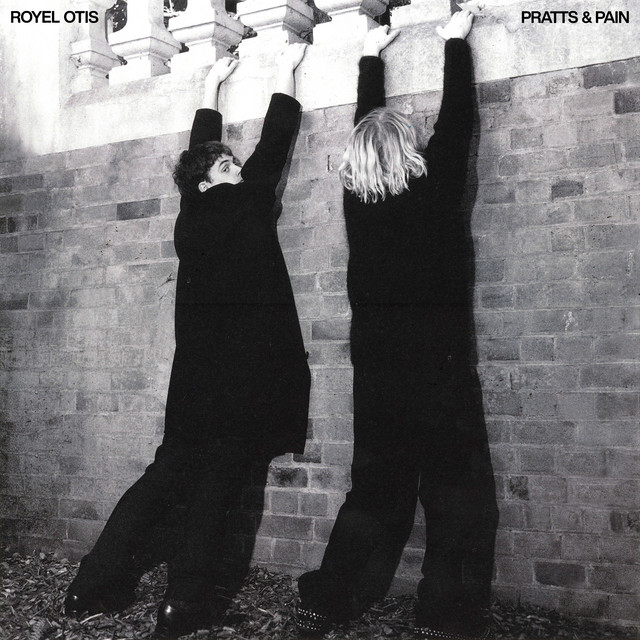 PRATTS & PAIN by Royel Otis album cover featuring two men dressed in black pretending to hang from a balcony by their hands. 