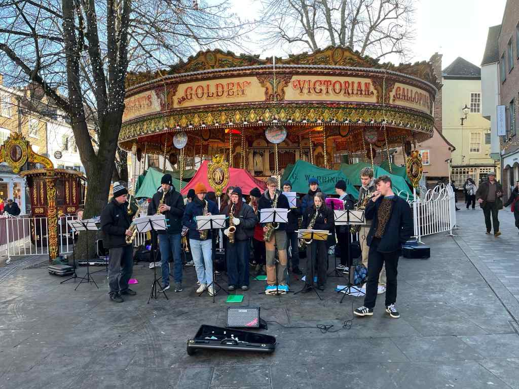 An image of around twenty students playing instruments with a lead male singer. They are playing on the streets in front of a carousel. 