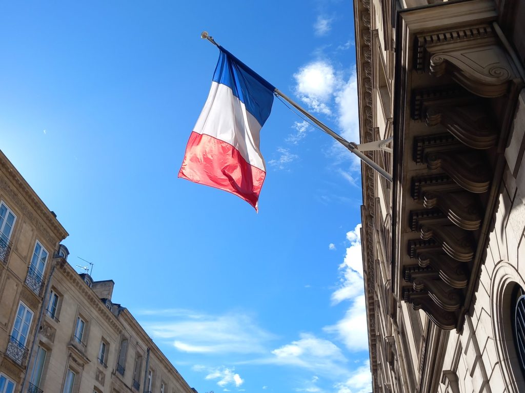 Photo of the French tricolour flag hanging on a pole jutting out from a buildling, with a blue sky in thebackground.