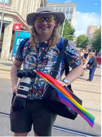 A photo of a person standing in touristy clothes, wearing a hat, sunglasses, hawaiian shirt, and holding a camera. they have an lgbt flag next to them