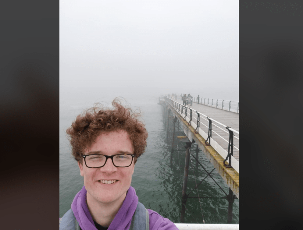 A photo of Harry Naylor. He is stood by a pier with the ocean descending into fog behind him. His ginger hair is windswept and he is smiling.