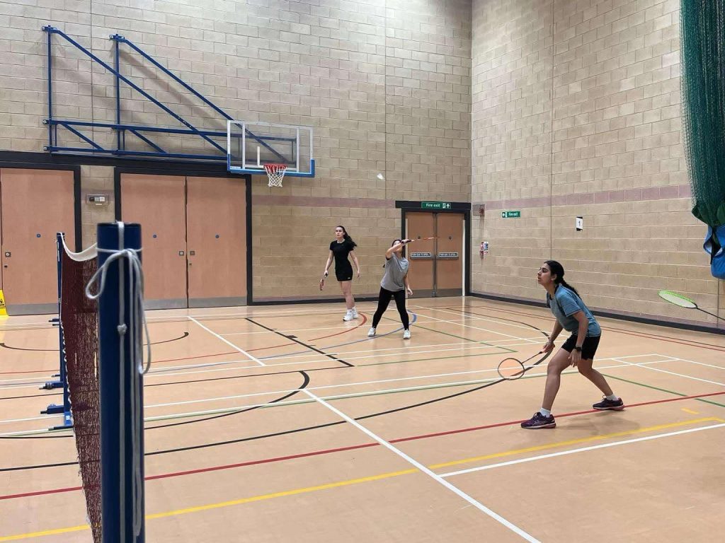 A photo of three girls playing badminton in a sports hall. You can see two courts and that they are playing two different games.