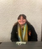 A photo of a girl smiling from a table, she is wearing a scarf and a trevs puffer. 
