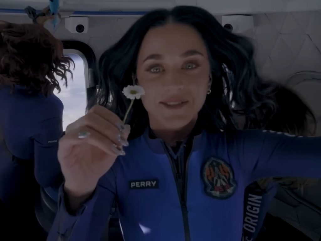 Katy Perry floats in a space capsule, she stares into the camera, holding a daisy flower, with the window to the capsule behind her.