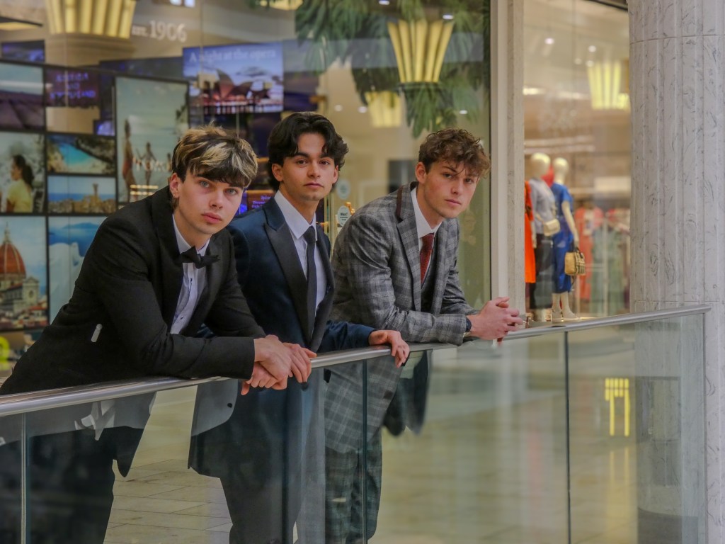 3 boys in suits lean over a railing at a shopping centre, all are looking at a camera