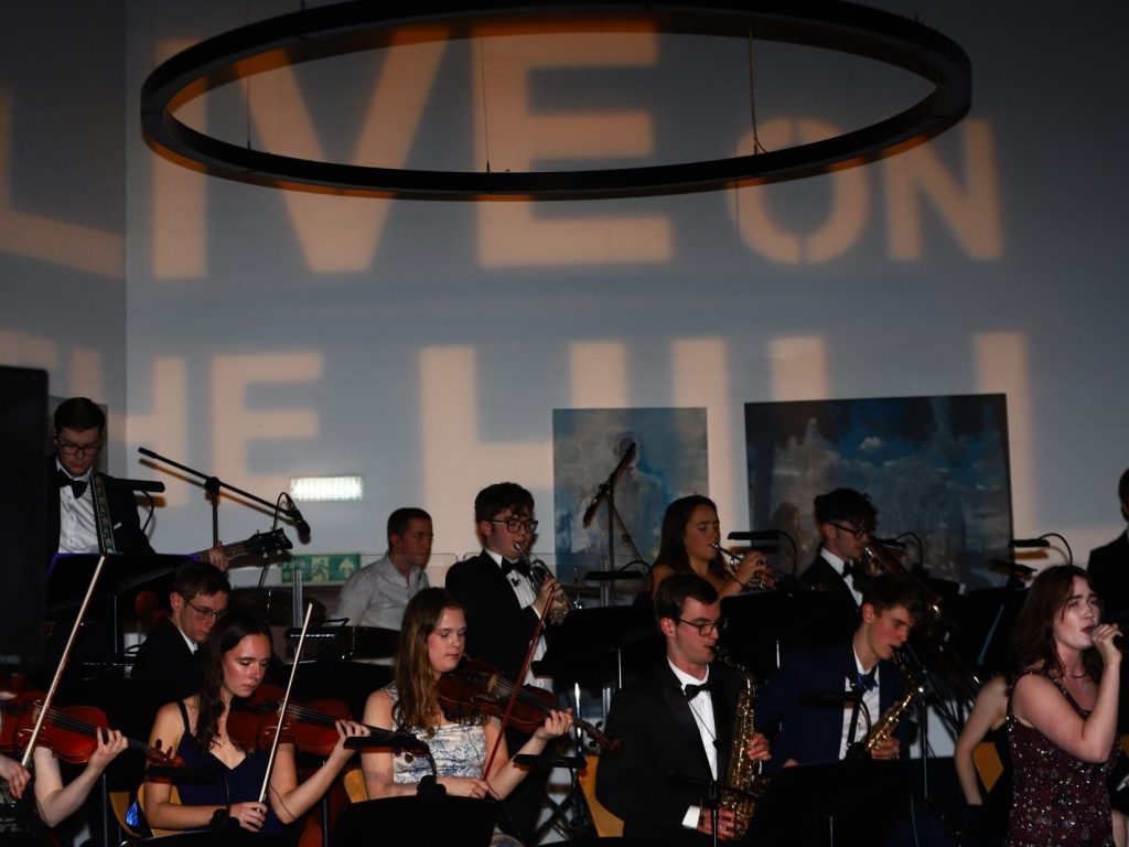 Orchestra and singer perform on stage with Live on the Hill in lights on wall behind them