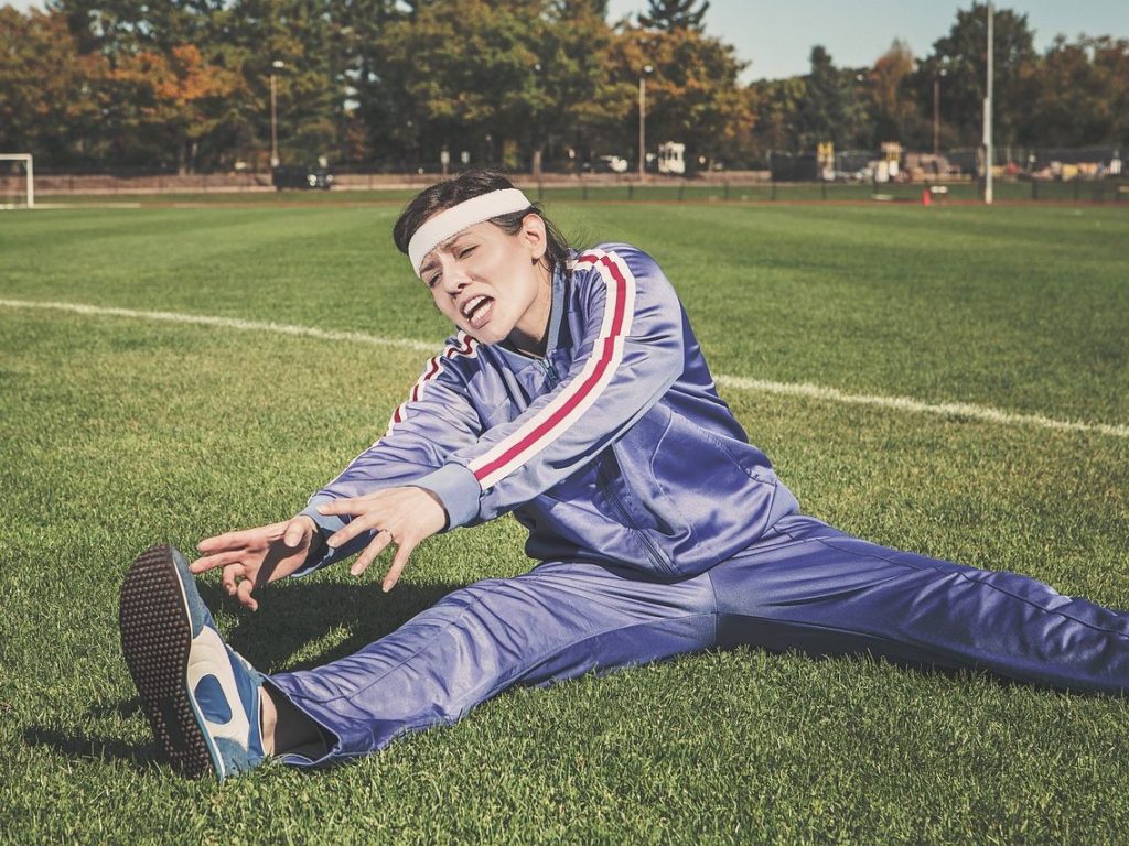 A women in a blue tracksuit and workout headband strains to stretch while sitting on a grass field.