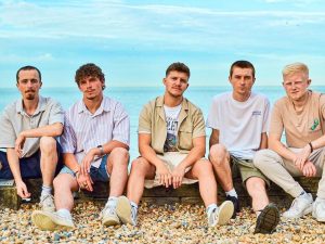 A picture of the five current members of Pool Club, sitting on a beach.